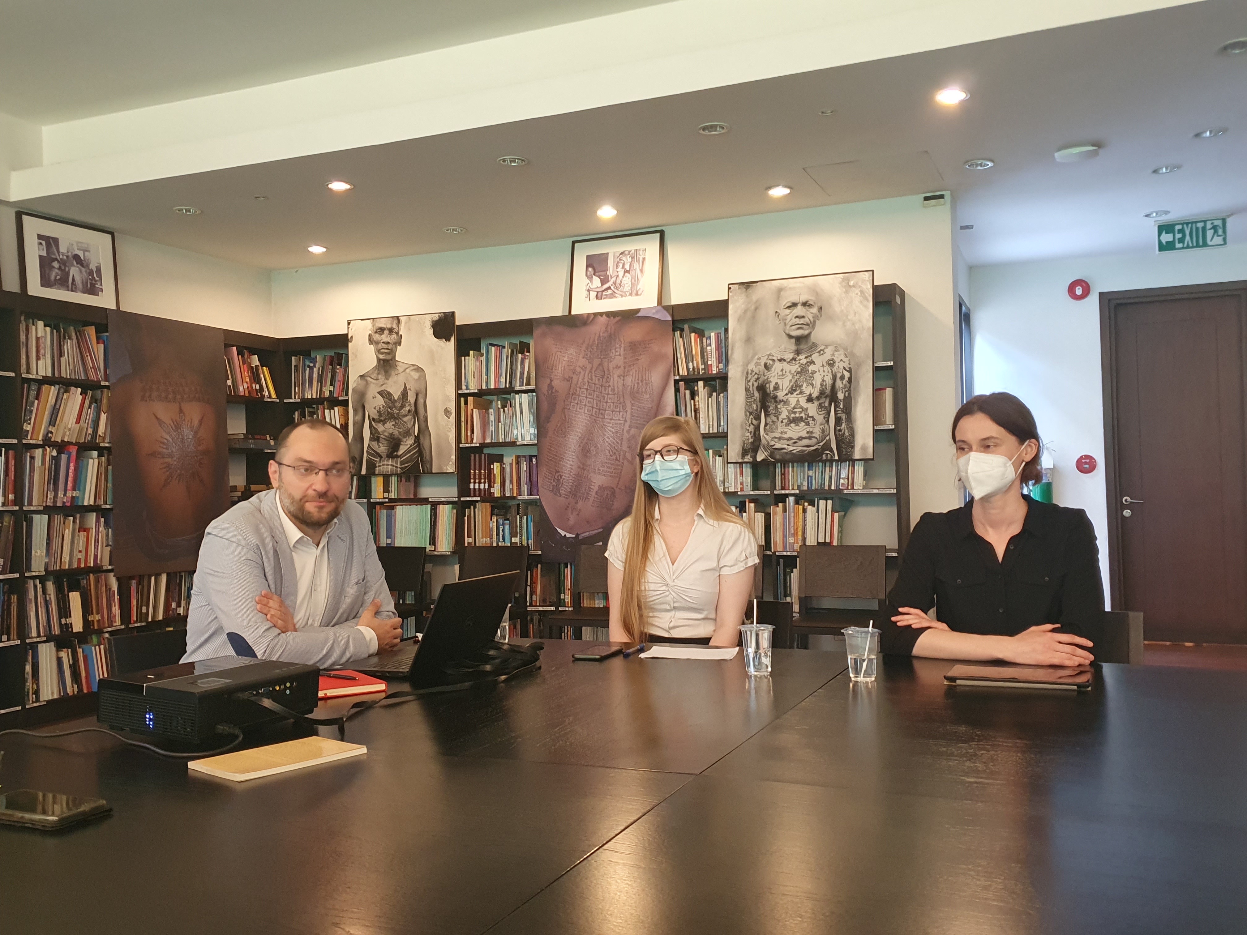 Jeden mężczyzna oraz dwie kobiety siedzą przy stole w pomieszczeniu z regałami, na których znajdują się książki/One man and two women are sitting at the table in the room with bookshelves on which there are books